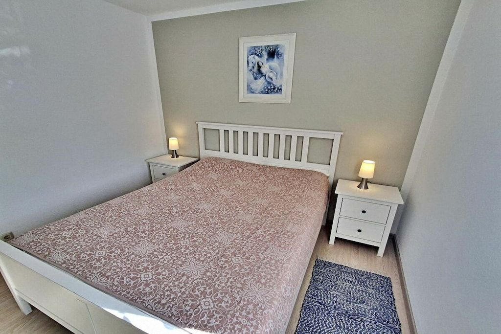 A bedroom with a bed, two bedside tables, a nightstand, a rug, and a wall-mounted picture.