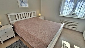 Une chambre à coucher comprenant un lit, une table de chevet, une lampe, une fenêtre avec des rideaux, un radiateur et un tableau encadré au mur.