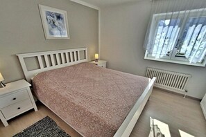 Un dormitorio con cama, mesita de noche, lámpara, ventana con cortinas, radiador y un cuadro enmarcado en la pared.
