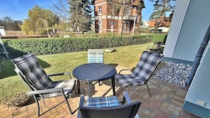 Une terrasse extérieure avec une table ronde et quatre chaises, une haie et un bâtiment en arrière-plan.