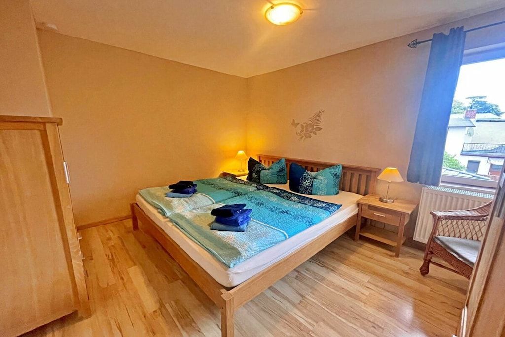 Un dormitorio con cama, mesita de noche, lámpara, silla y vistas al exterior.