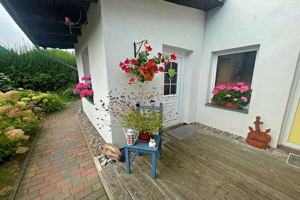 En träveranda med en krukväxt och ett litet bord, en vit dörr och ett fönster med rosa blommor.