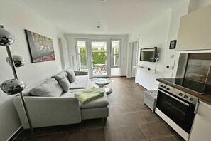 A modern living room with a sofa, a television, and a kitchen area with an oven. - Holiday apartment Altenberg 02 (Ahlbeck (Seebad))