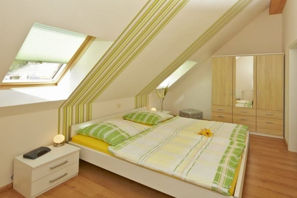 A bedroom with a sloped ceiling, a bed with a striped bedspread, a bedside table with a lamp, and a wardrobe.