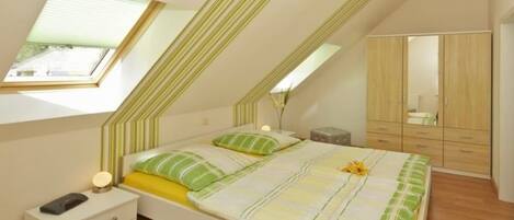 A bedroom with a sloped ceiling, a bed with a striped bedspread, a bedside table with a lamp, and a wardrobe.
