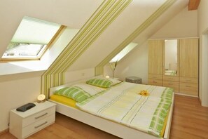 Quarto com teto inclinado, cama com roupa de cama listrada, mesa de cabeceira com abajur e guarda-roupa.