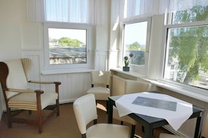 A room with a table, two chairs, and a window with a view of a street and greenery. - Holiday apartment with 1 bedroom (Ahlbeck (Seebad))