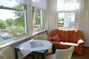 Une pièce comprenant une table, un fauteuil confortable et une fenêtre donnant sur un espace verdoyant et un bâtiment.