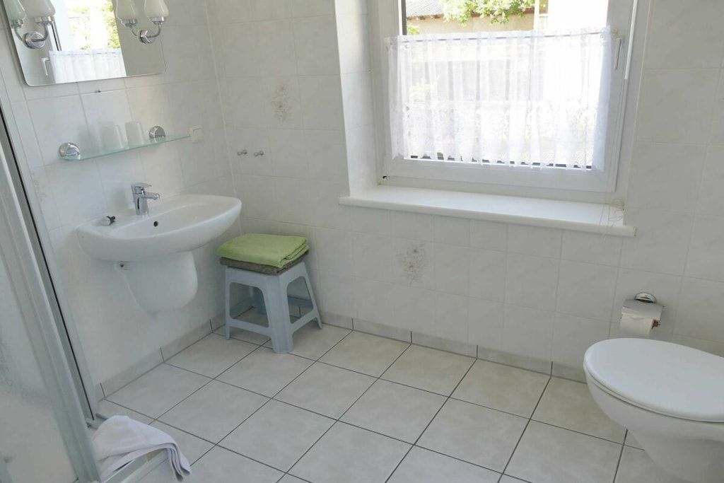 A bathroom with a white sink, a toilet, a small stool, and a window with a white blind.