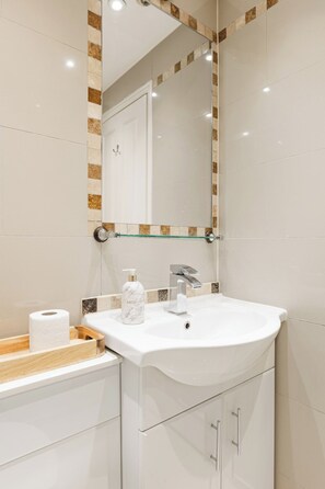 A modern bathroom with a white sink, a glass shelf, and a mirror with a decorative frame. - New! Spacious 1BR Flat | Marble Arch | Hyde Park (Greater London)