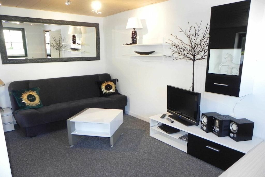 A modern living room with a sofa, a coffee table, a flat-screen TV, a TV stand with speakers, a lamp, and a mirror.