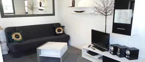 A modern living room with a sofa, a coffee table, a flat-screen TV, a TV stand with speakers, a lamp, and a mirror.