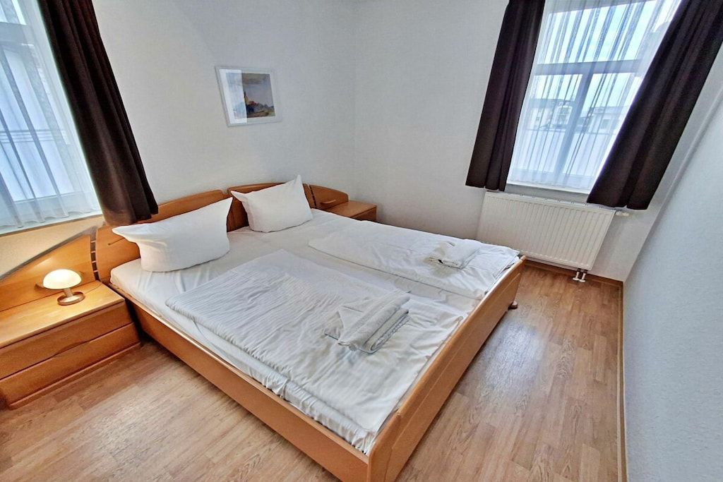 A neatly made double bed with white linens, a bedside table with a lamp, and a window with curtains.