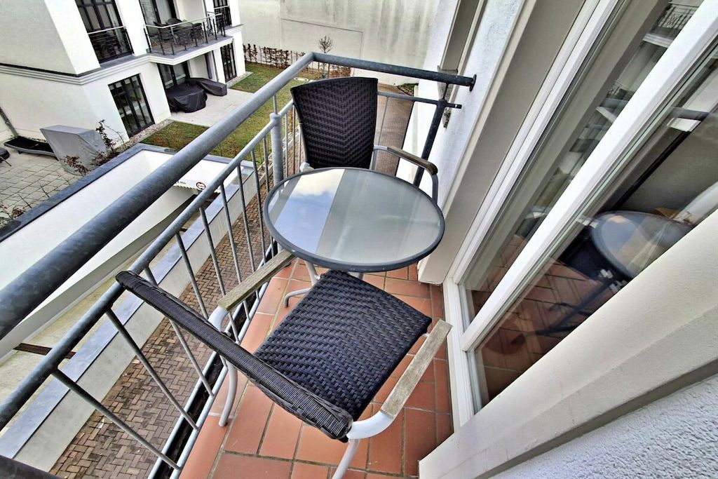 Un balcon avec une table en verre et des chaises en osier, donnant sur une zone pavée et un immeuble doté d’un balcon.