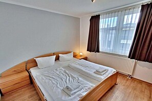 Una cama doble con sábanas y toallas blancas, un cabecero de madera, una mesita de noche con lámpara, una ventana con cortinas y un radiador.