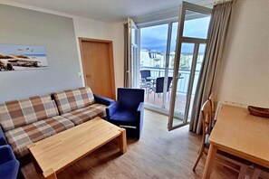 Sala de estar con sofá, sillón y mesa de centro. Una puerta da a un balcón exterior con mesa y sillas.