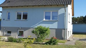 Una casa a due piani con tetto in tegole grigio, pareti bianche e un piccolo giardino anteriore con un albero e degli arbusti.