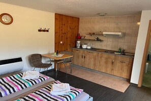 A compact room with a bed, a small table, a chair, a kitchenette, and a clock on the wall. - Feldmann holiday accommodations (Korswandt)