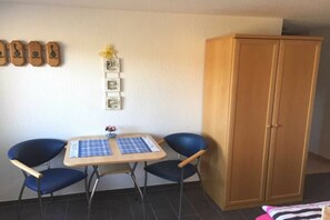 Quarto com guarda-roupa de madeira, uma mesa pequena com toalha xadrez, duas cadeiras azuis e uma parede com quadros.