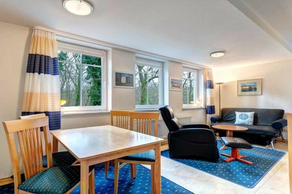 A room with a dining table, chairs, a sofa, and a small table with a lamp. There are windows with curtains, and a picture on the wall.
