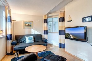 Ein kompakter Wohnraum mit einer Couch, einem kleinen runden Tisch, einem an der Wand befestigten Fernseher und einem Fenster mit Vorhängen.