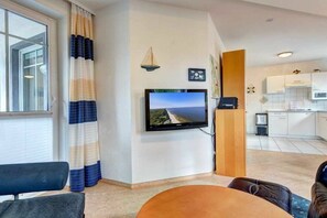 Ein Wohnzimmer mit an der Wand montiertem Fernseher, einem Esstisch und Blick auf die Küche im Hintergrund.