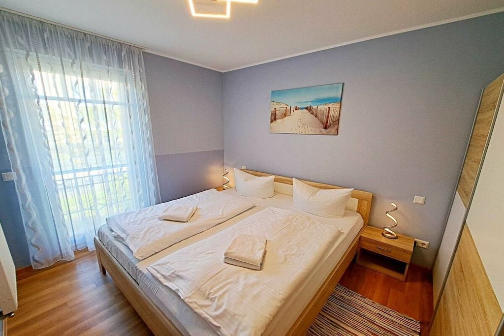 Ein Schlafzimmer mit einem Doppelbett, einem Nachttisch, einem Beistelltisch, einem Bild an der Wand und einem Fenster mit durchsichtigen Vorhängen.