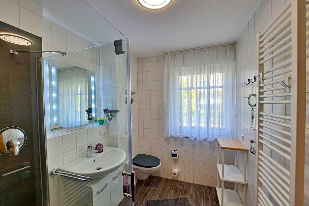 Ein Badezimmer mit einer gläsernen Duschkabine, einem Waschbecken mit Spiegel, einer Toilette und einem Fenster mit Vorhängen.