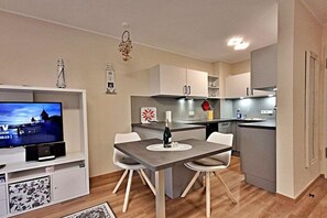 A modern living room with a flat-screen TV, a dining table with chairs, a kitchen area with cabinets and a stove, and a small decorative item on the wall. - Schloonseevillen Bansin Apartment 15 (Bansin (Seebad))