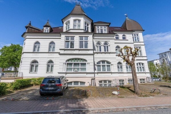 Un grand bâtiment blanc à plusieurs étages, doté d’une façade symétrique, d’une entrée centrale et d’un espace de stationnement où se trouve une voiture.