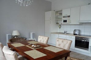 Un coin repas comprenant une table en bois, quatre chaises, un lustre, une lampe, ainsi qu’une cuisine avec des meubles et des appareils électroménagers blancs.