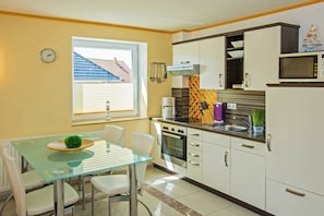 A modern kitchen with a glass dining table, white cabinets, and a window with blinds. - Holiday apartment with 1 bedroom (Heringsdorf (Seebad))