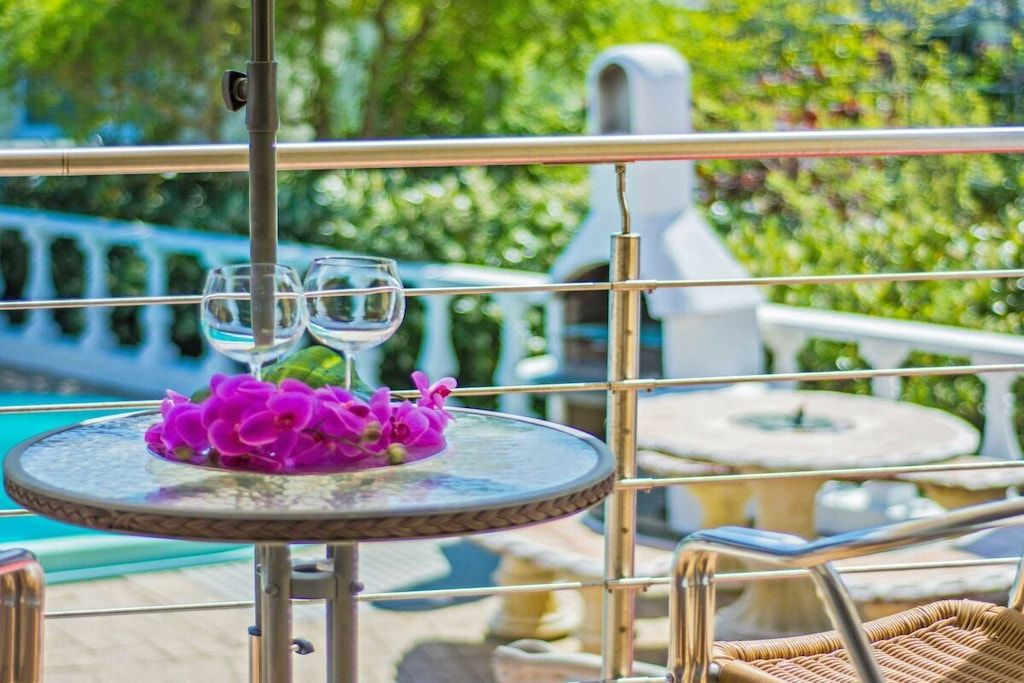 A balcony with a round table, two glasses, and purple flowers.