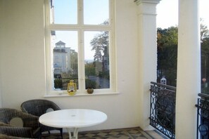 A room with a white table and wicker chairs, a window with a view of buildings, and a black wrought iron railing. - Villa Franz Josef 04 (Heringsdorf (Seebad))