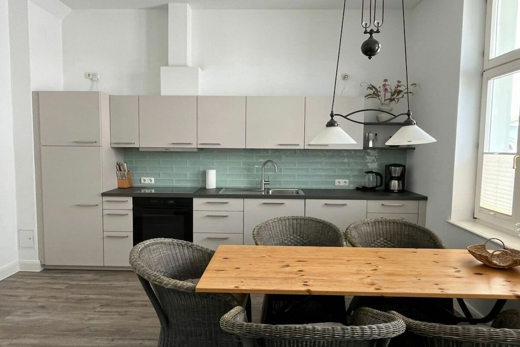 Une cuisine moderne avec des meubles blancs, une table à manger en bois, des chaises en osier et des luminaires suspendus.