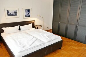 Cama bem arrumada com lençóis brancos em quarto com piso de madeira, cabeceira escura e armário fechado.