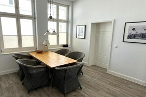 Uma área de refeições com mesa de madeira, cadeiras de vime, janelas amplas e quadros decorativos nas paredes.