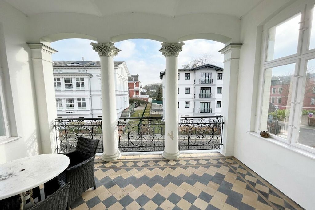 A balcony with a checkered floor, white columns, and a view of surrounding buildings.