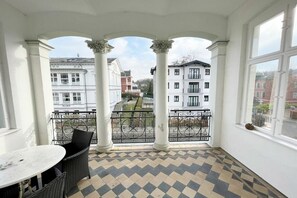 Varanda com piso xadrez, colunas brancas e vista para edifícios ao redor.