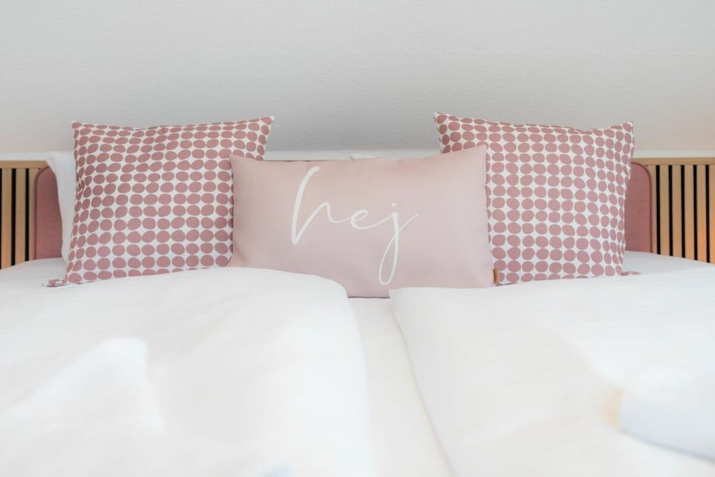 A bed with white bedding and pink polka dot pillows, one with the word 'hej' on it.