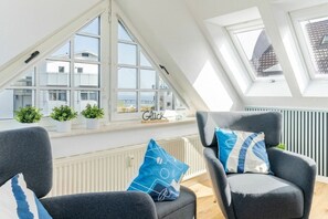 Ein Zimmer mit schrägen Wänden, großen Fenstern und zwei Sesseln mit Kissen.
