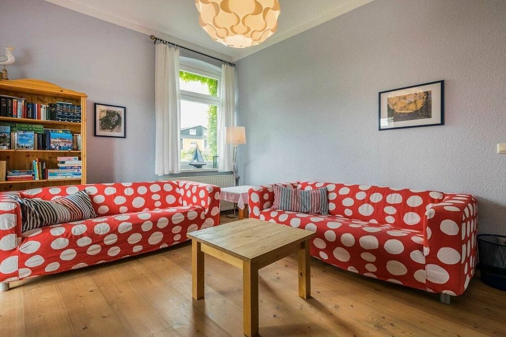 A living room with two red sofas with white polka dots, a wooden coffee table, a bookshelf, and a window with curtains.