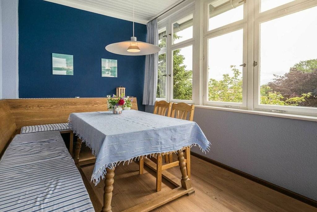 Une pièce avec un banc en bois, une table recouverte d’une nappe bleue et une fenêtre donnant sur un paysage verdoyant.