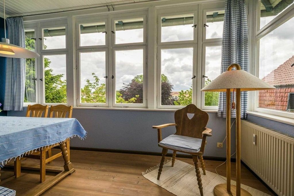A room with a dining table, chairs, a lamp, and large windows with curtains.