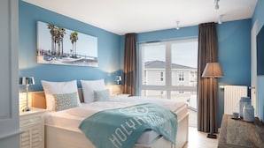 A bedroom with a large bed, a view of the sea, and a beach scene on the wall. - Vrbo Property (Heiligenhafen)