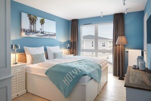 A bedroom with a large bed, a view of the sea, and a beach scene on the wall. - Vrbo Property (Heiligenhafen)