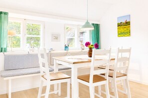 Ein helles Esszimmer mit einem Holztisch, weißen Stühlen, einer gestreiften Bank und einer hängenden Leuchte.