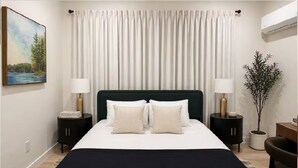 Quarto com uma cama, duas mesas de cabeceira, um criado-mudo, uma planta em vaso e um quadro na parede.