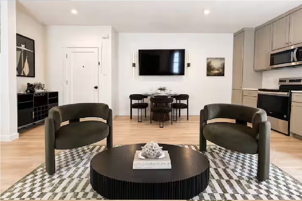 A modern living room with a round coffee table, two armchairs, a dining area with a table and chairs, a flat-screen TV mounted on the wall, and a kitchen area with cabinets and appliances.