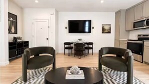 Uma sala de estar moderna com uma mesa de centro redonda, duas poltronas, uma área de jantar com mesa e cadeiras, uma TV de tela plana na parede e uma cozinha com armários e eletrodomésticos.